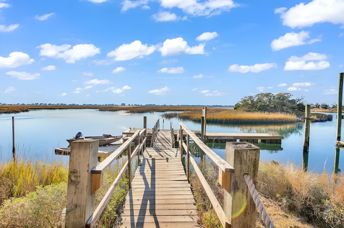 A wooden dock extends into calm waters, surrounded by tall grasses and scattered trees. The scene is illuminated by a clear blue sky with fluffy white clouds, enhancing the serene atmosphere of the marshland. A small boat is visible at the dock's edge.