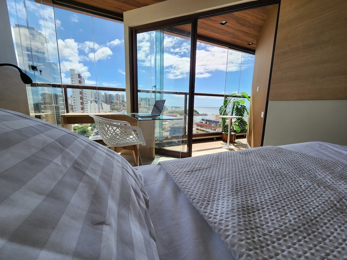 A spacious bedroom features large windows offering panoramic views of the city and sea. A comfortable bed is adorned with a textured coverlet. A small workspace with a laptop rests beside the window, allowing for natural light to fill the room.