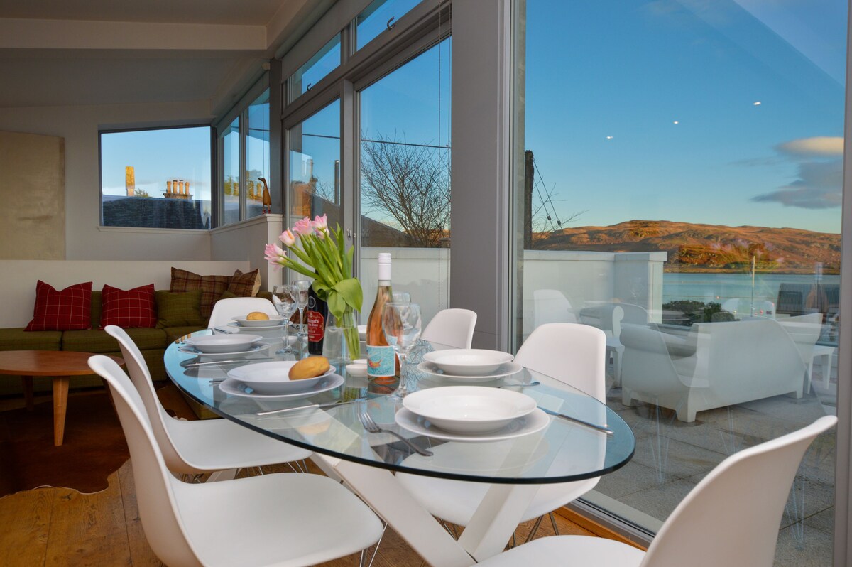 A modern dining area features a glass table surrounded by white chairs. Fresh flowers are placed in the center, while table settings include plates and glasses. Expansive windows offer views of the surrounding landscape and create a bright atmosphere.
