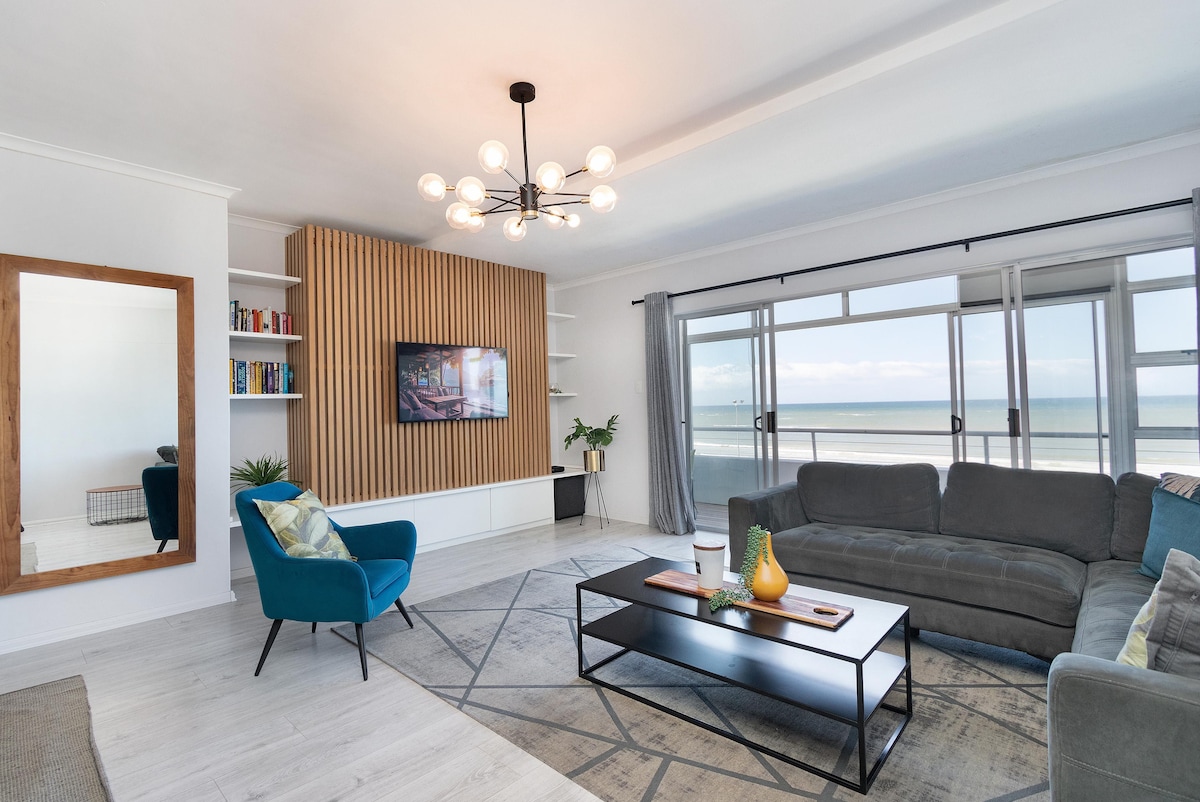A modern lounge area features a large grey sectional sofa and a coffee table with decorative items. A wall-mounted television is positioned above a shallow storage unit with books. Large windows overlook the sea, allowing natural light to fill the space. A tall mirror adds depth to the room.
