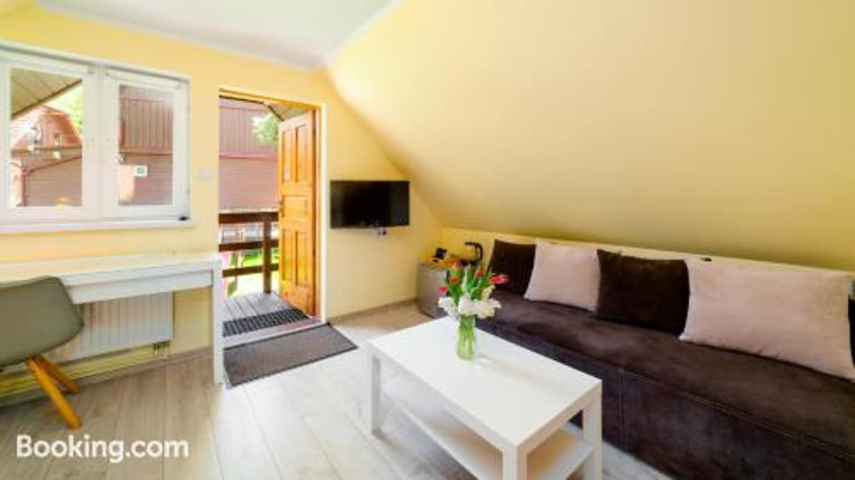 A bright sitting area features a brown sofa with cushions, paired with a small white coffee table. A television is mounted on the wall, while a desk and chair provide workspace. Natural light enters through the window and door, enhancing the warm yellow walls.