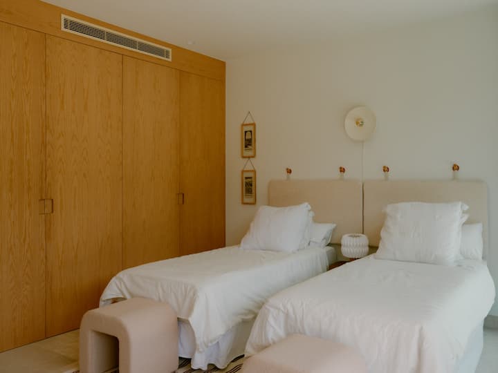 Una habitación grande con dos camas individuales cómodas, mucho espacio para guardar ropa y un espejo de cuerpo entero. La habitación incluye una silla y un banco para mayor comodidad. También se abre a una terraza privada amueblada con dos sillas y una mesa de café