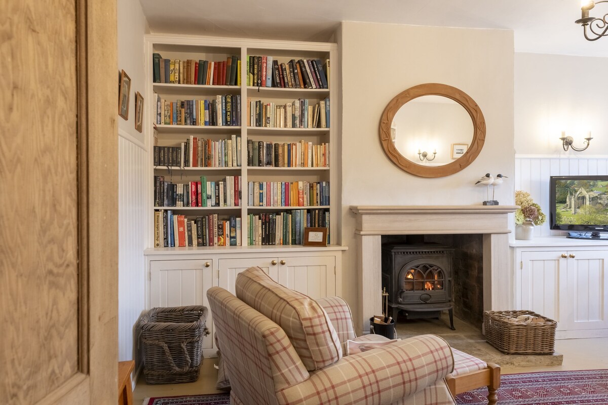 A cozy sitting area features a stone fireplace with a wood-burning stove, surrounded by light-colored cabinetry. A bookshelf filled with various books is positioned against the wall. A comfortable sofa sits invitingly, complemented by a classic round mirror above the fireplace.