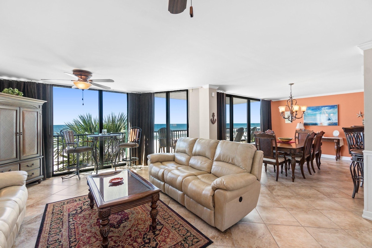 A spacious living area features large floor-to-ceiling windows that provide views of the beach and ocean. Comfortable seating is arranged around a decorative coffee table, and a dining area with a wooden table is visible in the background. Ocean-themed artwork adorns the walls.