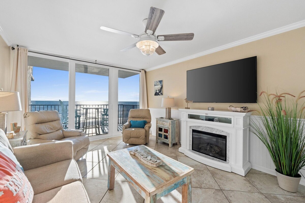 The living area is equipped with a comfortable sofa and two reclining chairs, facing a modern television mounted above a decorative electric fireplace. Large glass doors provide access to a balcony, allowing for views of the beachfront. Natural light fills the space, highlighting the coastal decor.