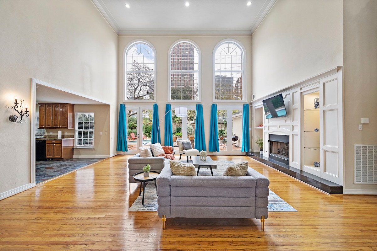 The spacious living room is characterized by high ceilings and ample natural light, provided by large arched windows. A pair of comfortable sofas is arranged around a coffee table. Vibrantly colored curtains frame the windows, enhancing the elegant ambiance.