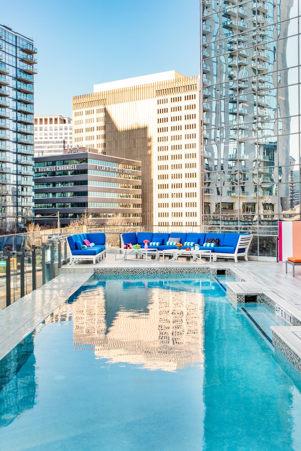 A vibrant rooftop pool area is featured, surrounded by sleek modern furniture in blue and white. The pool's clear water reflects the towering buildings of the cityscape, enhancing the tranquil environment. Glass railings provide an unobstructed view of the skyline.