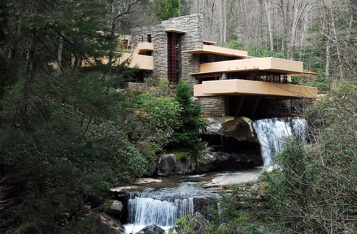 A modern architectural structure blends into a natural setting, surrounded by trees and boulders. Water cascades down from a rocky area, creating a serene environment. The building features multiple levels with horizontal lines and large windows reflecting the surrounding greenery.