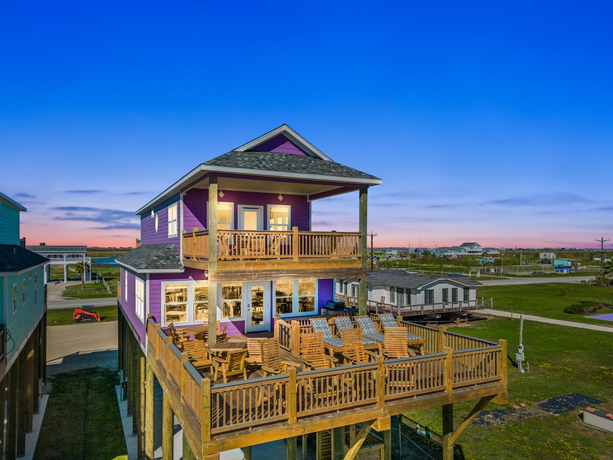 The exterior view showcases a multi-level beach house, featuring a prominent wooden deck with multiple chairs. The vibrant colors of the house contrast with the twilight sky, enhancing the coastal ambiance. Surrounding landscape presents open space and neighboring structures, contributing to the serene atmosphere.