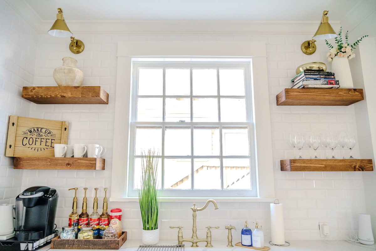 A cozy kitchen corner is highlighted by white subway tiles and soft natural light. Open wooden shelves showcase decorative items, glassware, and coffee essentials. A stylish coffee maker sits alongside containers, with a potted plant adding a touch of greenery to the space.