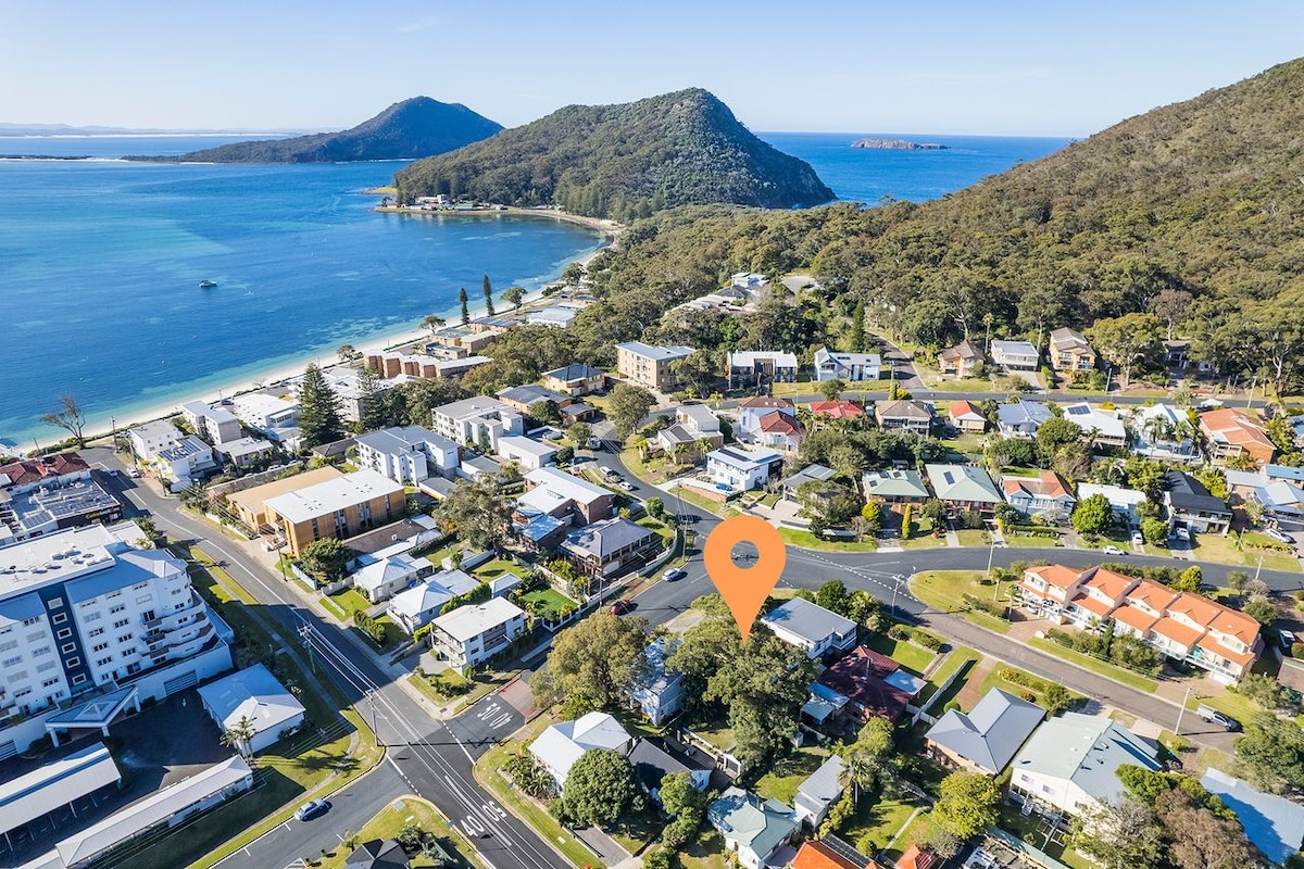 An aerial view showcases the Starfish Lodge's location within Shoal Bay. The surrounding area features a mix of residential buildings, green trees, and a glimpse of the tranquil blue waters of the bay, with rolling hills visible in the background.