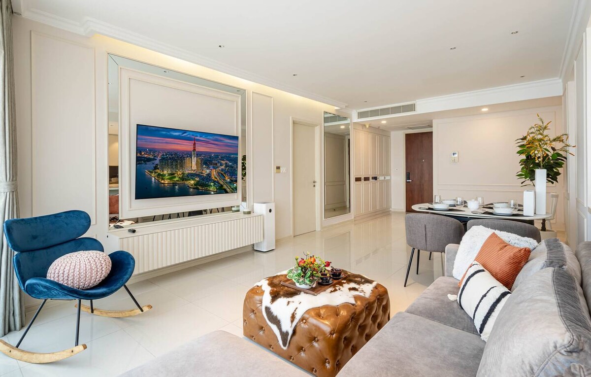 A modern living area showcases a neutral colour palette with elegant furnishings. A plush sofa is paired with a stylish rocking chair. A large flat-screen TV is mounted on the wall, reflecting a view of the city skyline. A dining area is set for four, with tasteful decor elements accenting the space.