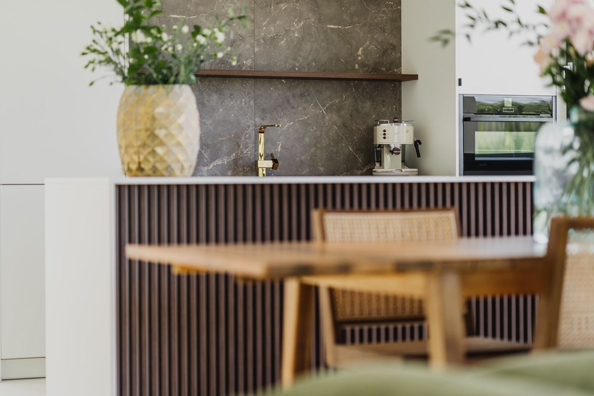 A modern kitchen area features a stylish countertop with a textured stone backsplash. A decorative vase filled with greenery sits prominently, while a coffee machine is positioned nearby. A wooden dining table with woven chairs complements the space.