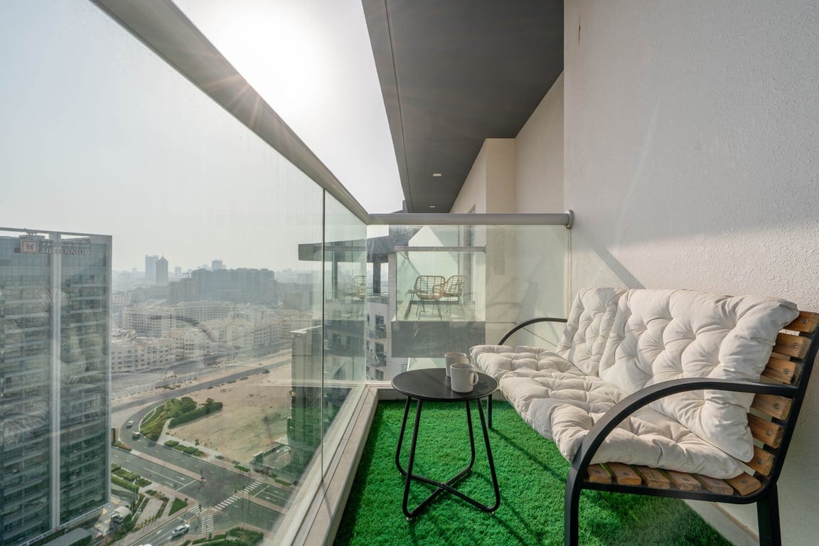 A comfortable balcony space is featured with a plush, cushioned bench and a small table. The lush green carpet adds a refreshing touch, while the view showcases the surrounding urban landscape, allowing for relaxation and enjoyment of the outdoors.
