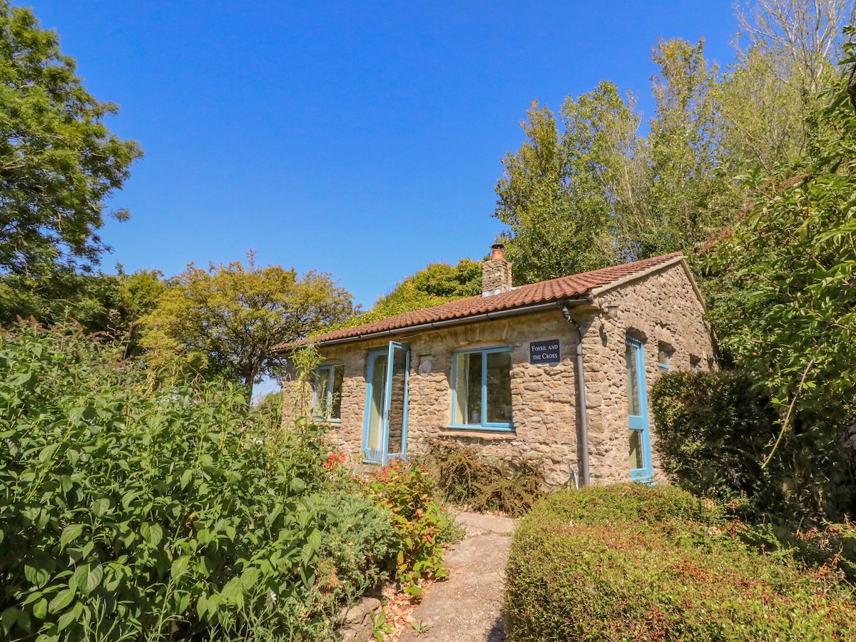 A charming stone cottage is surrounded by greenery, with clear blue skies overhead. Large windows frame the entrance, inviting natural light inside. A pathway leads to the door, flanked by well-maintained shrubs and plants that enhance the cottage's natural setting.
