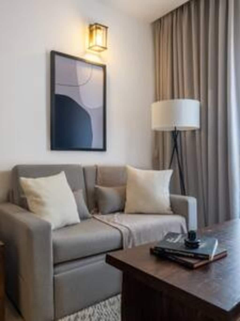 A cozy living area features a soft two-seater sofa adorned with beige cushions. A stylish lamp stands beside the sofa, complementing a modern wall art piece. A wooden table is positioned in front, showcasing a book and a decorative object.