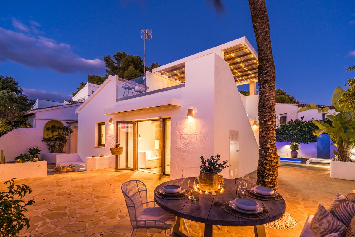 The exterior of the villa is presented in the twilight, showcasing smooth white walls and a terrace illuminated by soft lighting. Outdoor seating and a dining table create a welcoming space. Lush greenery frames the property, while the serene atmosphere hints at relaxation.