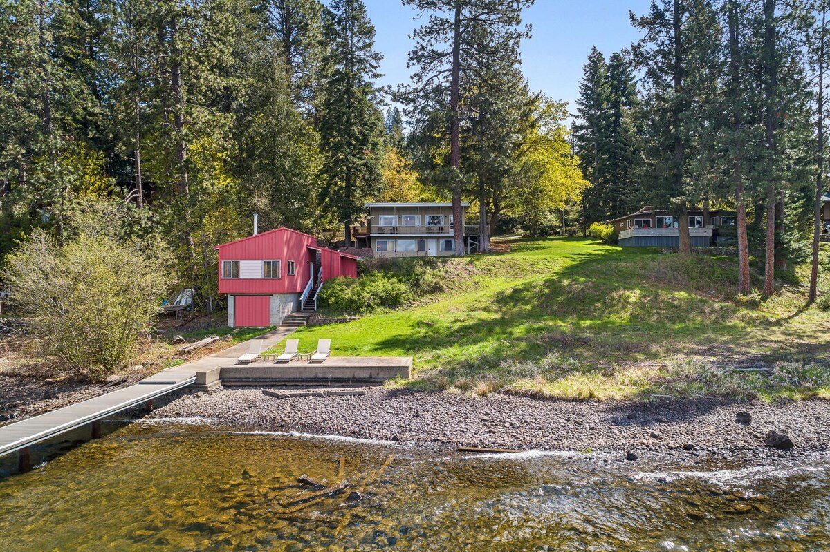 A lakefront view features a vintage cabin in red and a two-story structure, surrounded by lush greenery. A private dock with lounge chairs sits against a pebbled shore, providing easy access to the clear waters of the lake.