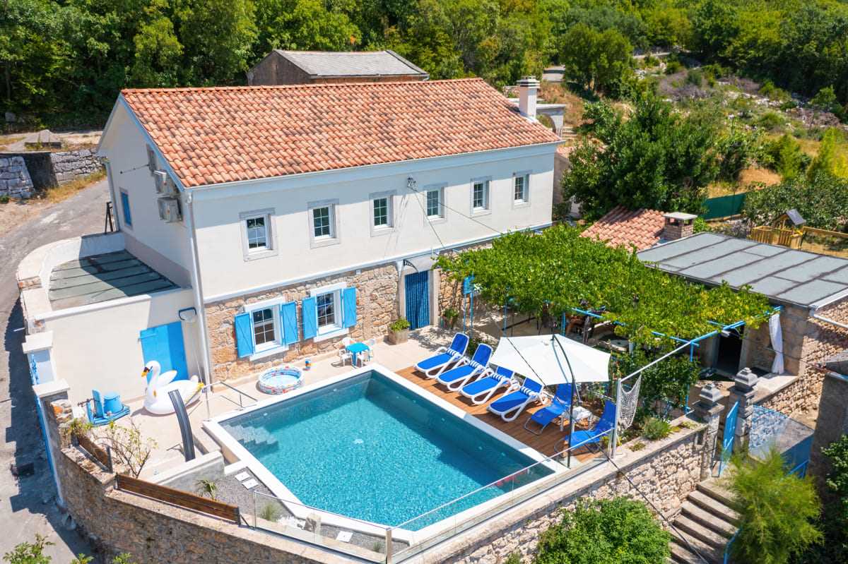 An aerial view of a two-story stone house features a well-maintained pool surrounded by sun loungers. A shaded seating area and outdoor vines contribute to the inviting outdoor space. Lush greenery landscape encompasses the property, enhancing the serene atmosphere.