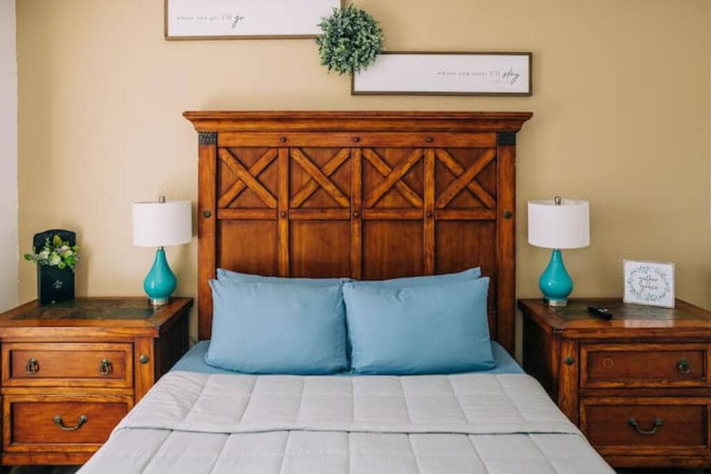 A wooden bed frame with an intricately designed headboard is centered against a beige wall. Two bedside tables, each adorned with a blue lamp and decorative items, flank the bed. Soft blue pillows are placed against a light-colored blanket on the bed.
