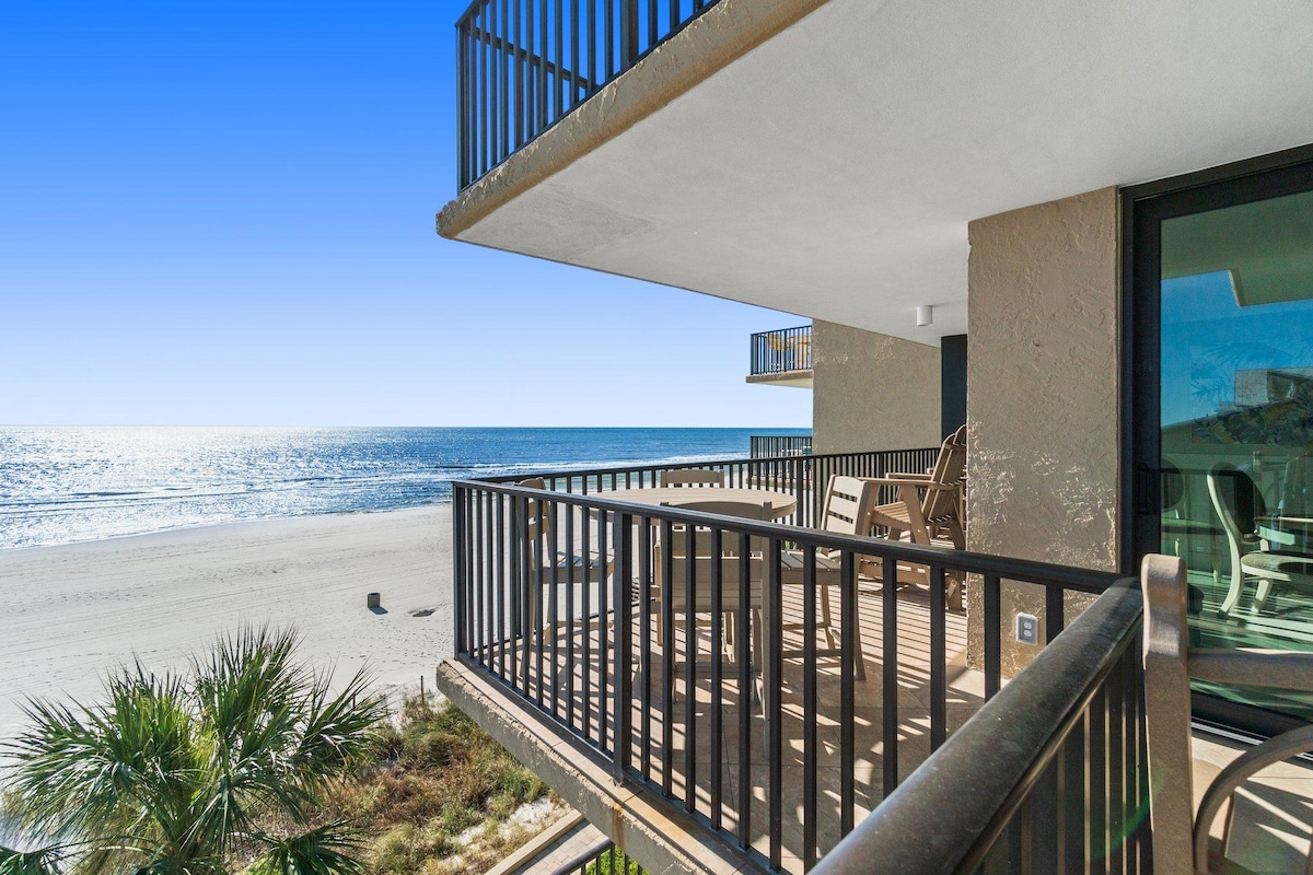 A corner balcony offers a clear view of the beach and Gulf of Mexico. Outdoor furniture is arranged near the railing, featuring a dining table and seating. The coastline and water reflect sunlight, creating a serene atmosphere.