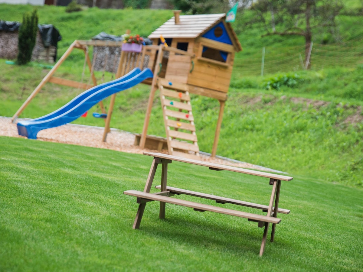 A wooden picnic table is situated on a well-manicured grassy area. In the background, a colorful wooden play structure is visible, featuring a slide and climbing steps, providing an inviting space for outdoor play.