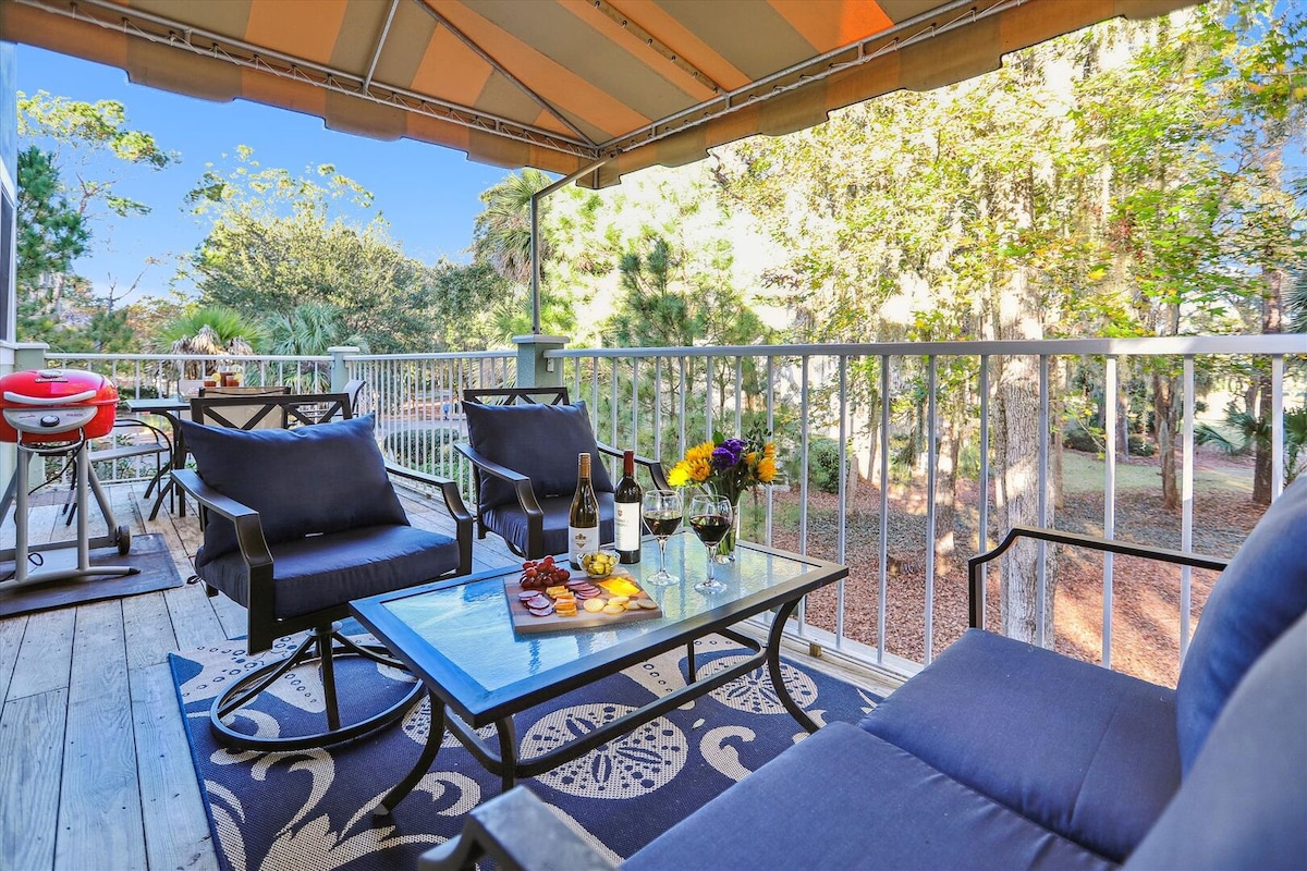 An expansive back deck features comfortable seating arranged around a glass table, ready for relaxation. A grill is visible, and a serene backdrop of natural greenery enhances the outdoor space, inviting guests to enjoy al fresco dining and leisure.
