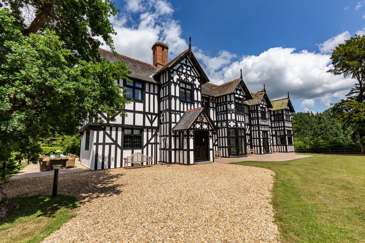 A traditional style house with a timber-framed exterior is set amongst an expansive lawn. The structure features multiple gabled rooftops and large windows, allowing natural light to illuminate the interiors. A gravel pathway leads to the entrance, enhancing accessibility and charm.
