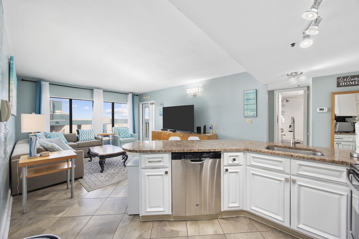 An open-concept living area features large windows allowing natural light to fill the space. A comfortable sofa is positioned beside a modern entertainment unit with a flat-screen TV. The kitchen showcases updated stainless steel appliances and a bar counter, enhancing the functional layout.