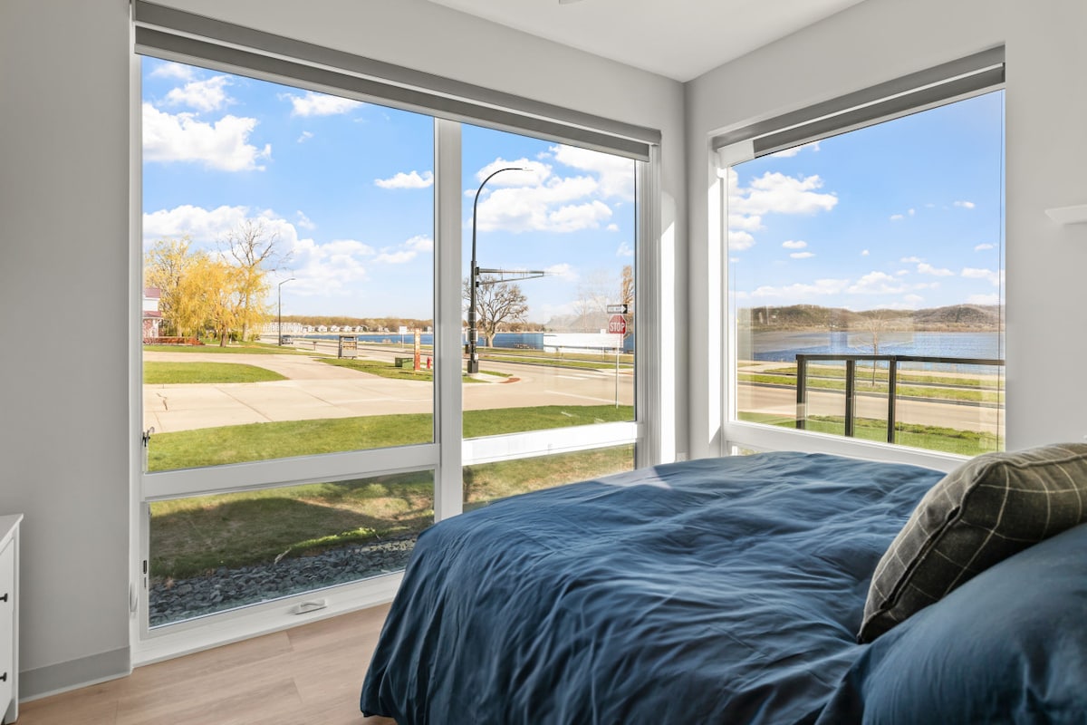 A corner bedroom features large windows providing views of the serene waters of Lake Pepin. Natural light fills the space, highlighting a spacious bed covered in dark bedding. Inviting blue skies and distant greenery enhance the tranquil setting outside.