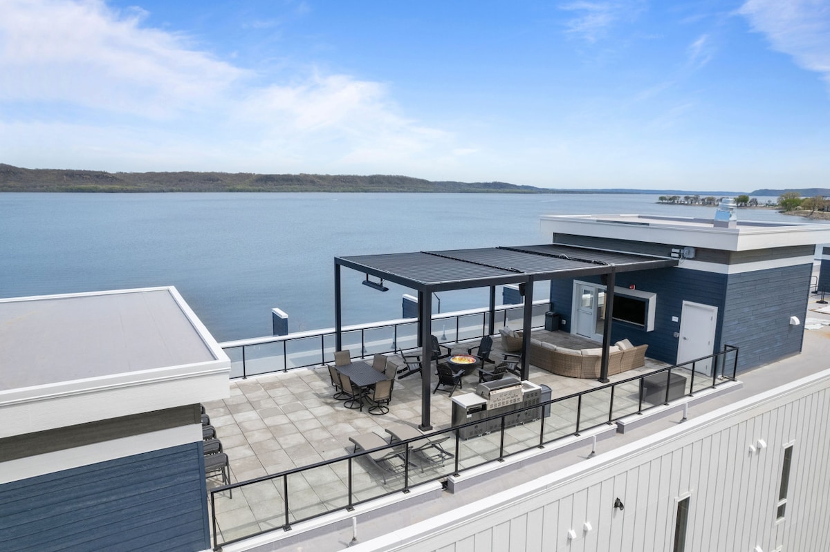 A rooftop terrace offers panoramic views of Lake Pepin. The outdoor space features a shaded area with seating arrangements and a hot tub, surrounded by a spacious patio. The serene lake reflects the clear sky, creating a calming atmosphere for gatherings.