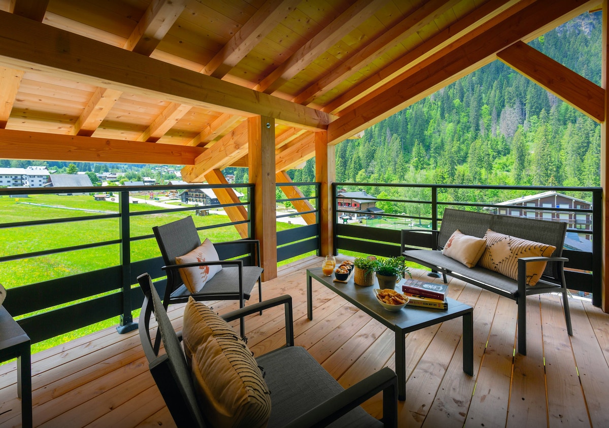 A spacious balcony features a wooden ceiling and floor, offering a view of surrounding green landscapes and the village below. Comfortable seating is provided by gray chairs and a coffee table adorned with decorative plants and books.