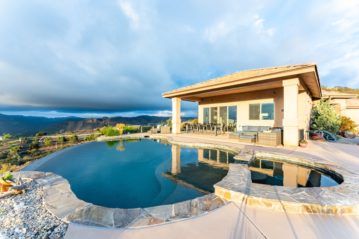 An elegantly designed outdoor space features a sparkling infinity pool with a hot tub. Stone accents complement the modern architecture of the house, which boasts large windows offering sweeping views of the surrounding mountains and sky.