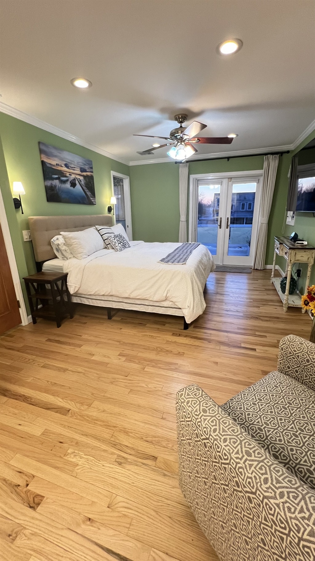 A spacious bedroom features a king bed dressed in white bedding, complemented by a cozy armchair in the corner. Large glass doors provide access to a sunlit outdoor area, while soft green walls and wood flooring enhance the room's inviting ambiance.