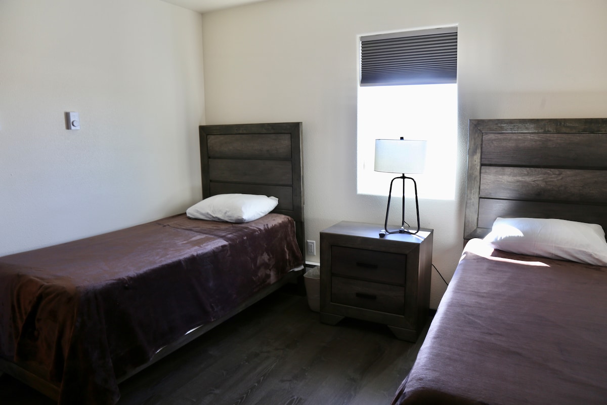 A well-lit bedroom features two twin beds, each with a neutral-toned blanket and a single white pillow. A nightstand with a lamp is positioned between the beds, and natural light enters through a window with light-filtering shades.