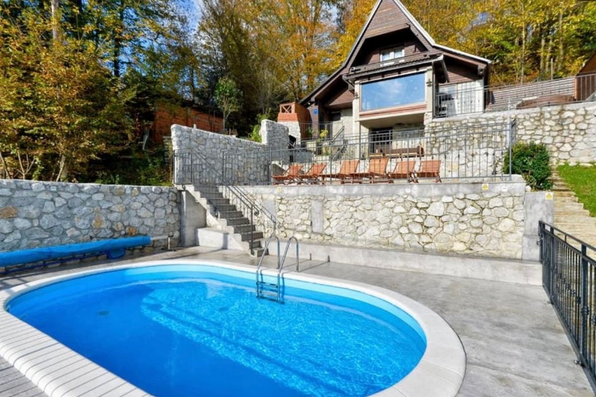 A private swimming pool is set against a stone wall, with steps leading up to a wooden terrace equipped with sun loungers. The surrounding area features greenery and trees, creating a serene atmosphere. A charming house with large windows is visible in the background.