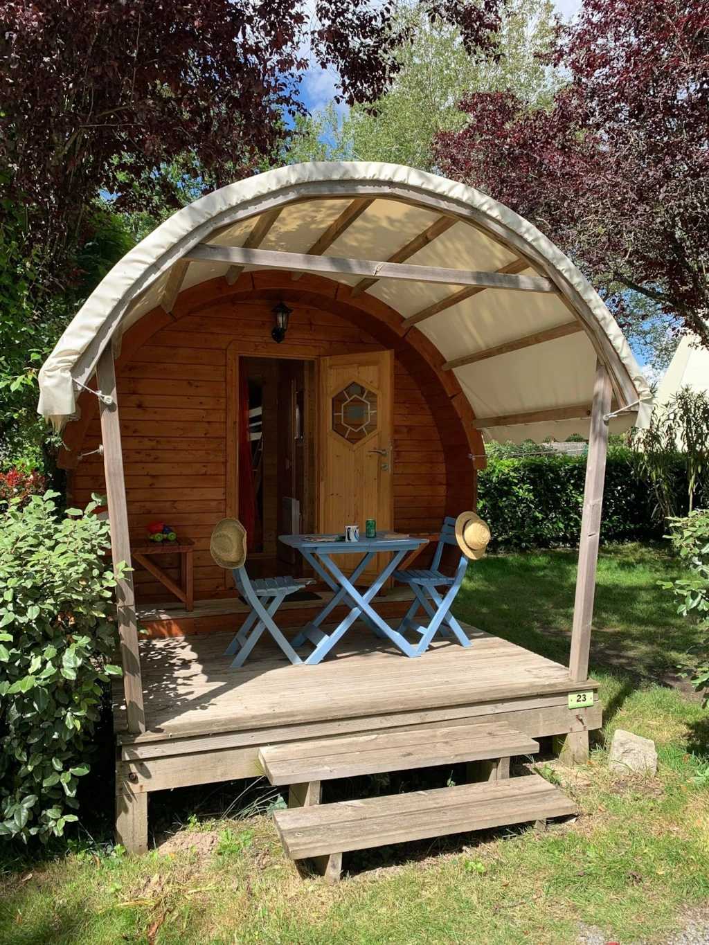 A charming wooden cabin features a curved roof and a welcoming entrance. Outside, a small porch is furnished with a blue table and two chairs, surrounded by greenery. The natural light creates a warm atmosphere, inviting guests to relax in the serene outdoor space.