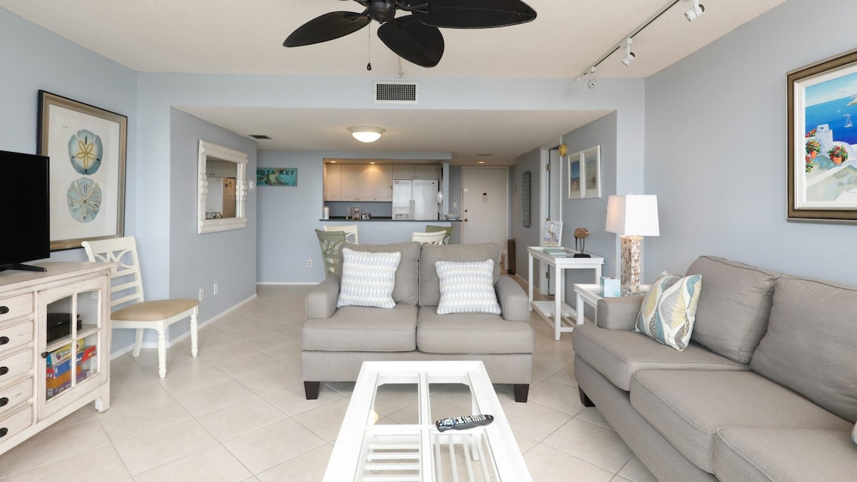 The spacious living area features soft grey furnishings, including a sectional sofa and a coordinating armchair. A glass coffee table sits in the center, and light walls complement the decorative art. The open layout connects to the kitchen, visible in the background.