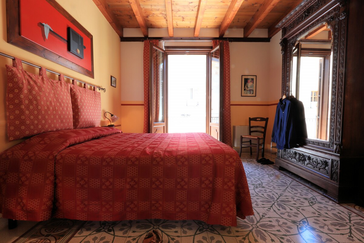 A room features a spacious bed covered in a rich red bedspread. Natural light enters through two windows framed by patterned curtains. A large, ornate mirror and a wooden chair complement the decor, while a jacket hangs casually on the mirror's edge.