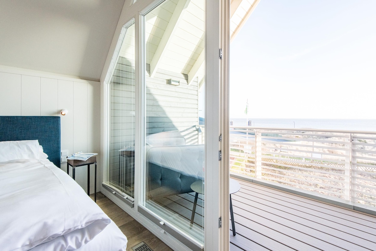 A spacious bedroom features large glass doors that open to a private balcony, showcasing views of the water. The elegant bed is adorned with crisp white linens, and a small table is positioned beside it, inviting relaxation amidst natural light.