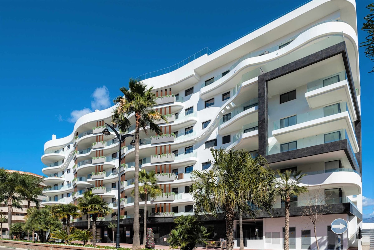 A modern building constructed with an elegant, wavy facade is surrounded by lush palm trees and landscaping. The structure features multiple balconies and large windows, reflecting a bright blue sky. The contemporary design reveals a spacious and inviting atmosphere.