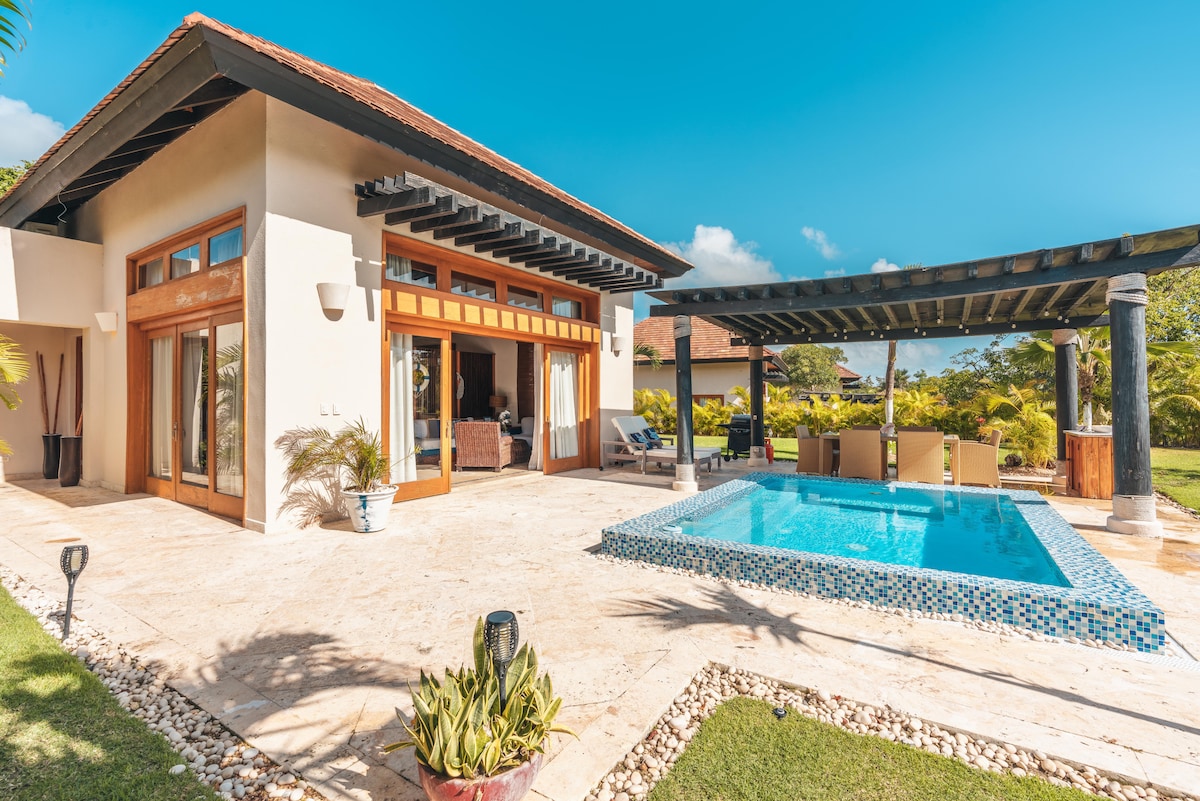 A private outdoor space is showcased, featuring a swimming pool with a blue-tiled edge. Comfortable seating is arranged under a covered patio. Large glass doors provide access to the interior, surrounded by well-maintained greenery and a stone pathway.