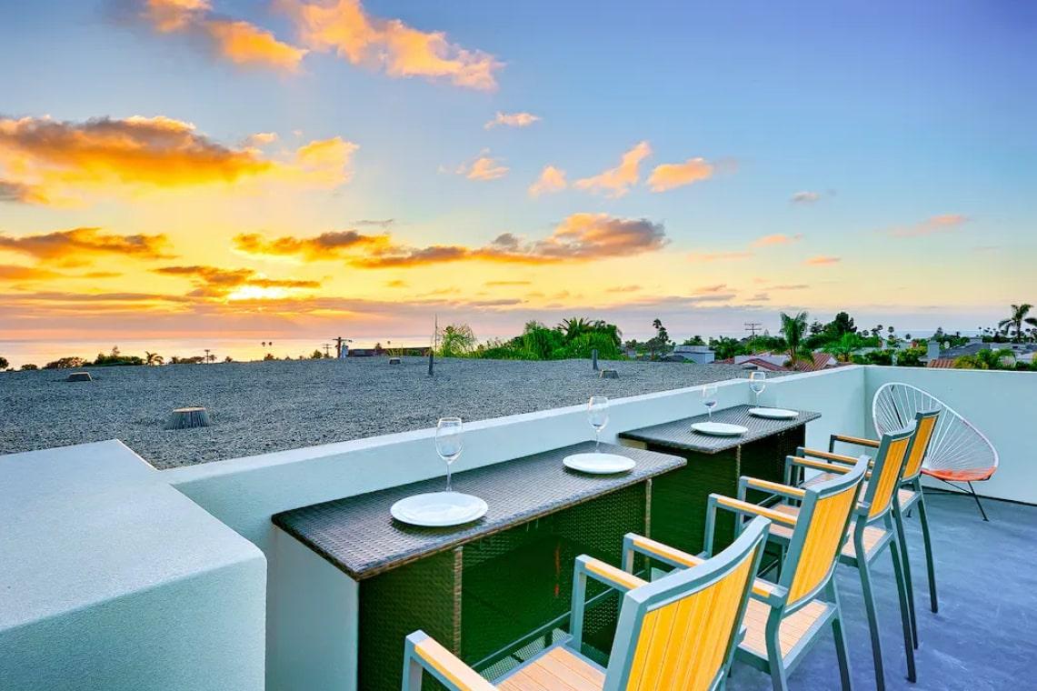 The rooftop deck features several high-top chairs positioned around a long bar overlooking the ocean. A vibrant sunset casts warm hues across the sky, enhancing the serene outdoor setting. Glasses are arranged on the bar, ready for guests to enjoy the view.