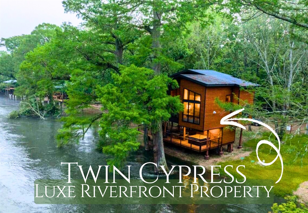 The cabin is showcased from an elevated perspective, surrounded by lush greenery and positioned adjacent to the river. A wooden deck extends from the structure, providing direct access to the serene waters. Large windows reflect the tranquil environment, inviting natural light indoors.