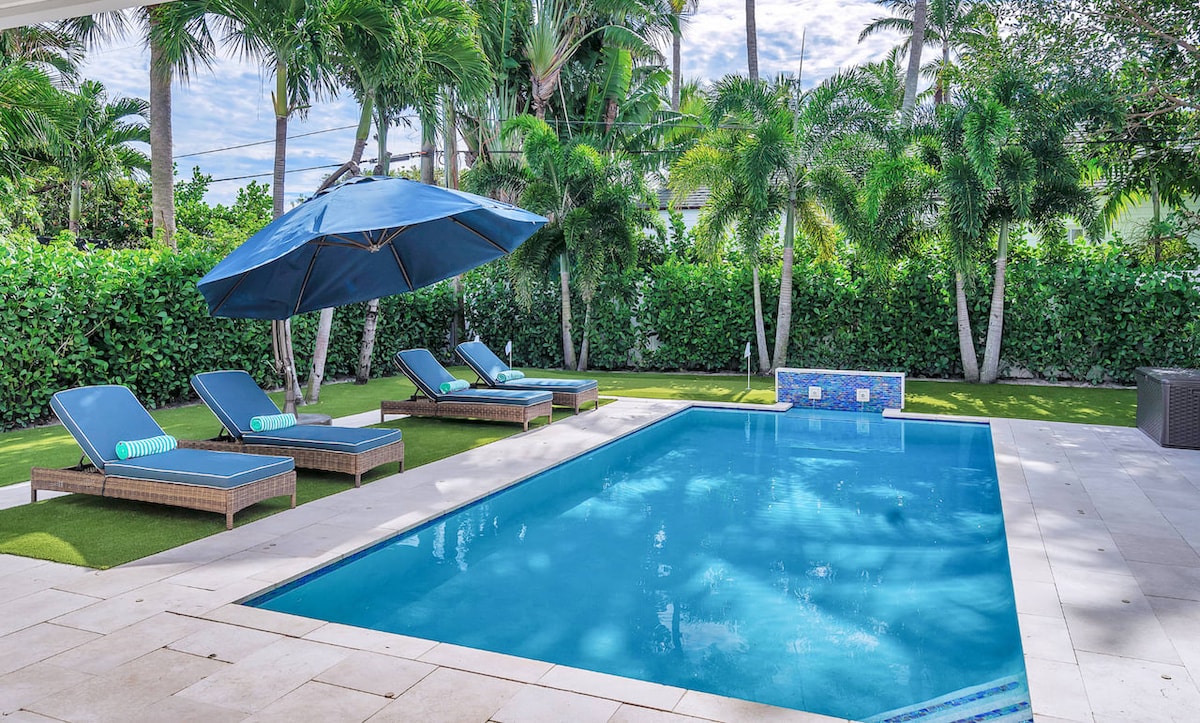 A serene backyard features a heated pool surrounded by lush greenery. Four lounge chairs with cushioned seating are positioned nearby, with a large blue umbrella providing shade. A mosaic-tiled seating area is seen at one end of the pool, promoting relaxation and enjoyment.