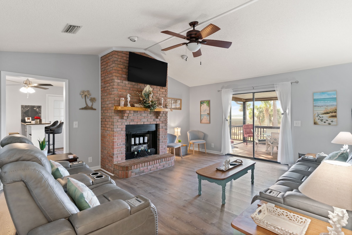 A spacious living area features a brick fireplace as a focal point, complemented by a ceiling fan and comfortable seating. Large windows provide a view of the outside, with sliding doors leading to a balcony. Decorative art pieces enhance the cozy atmosphere.