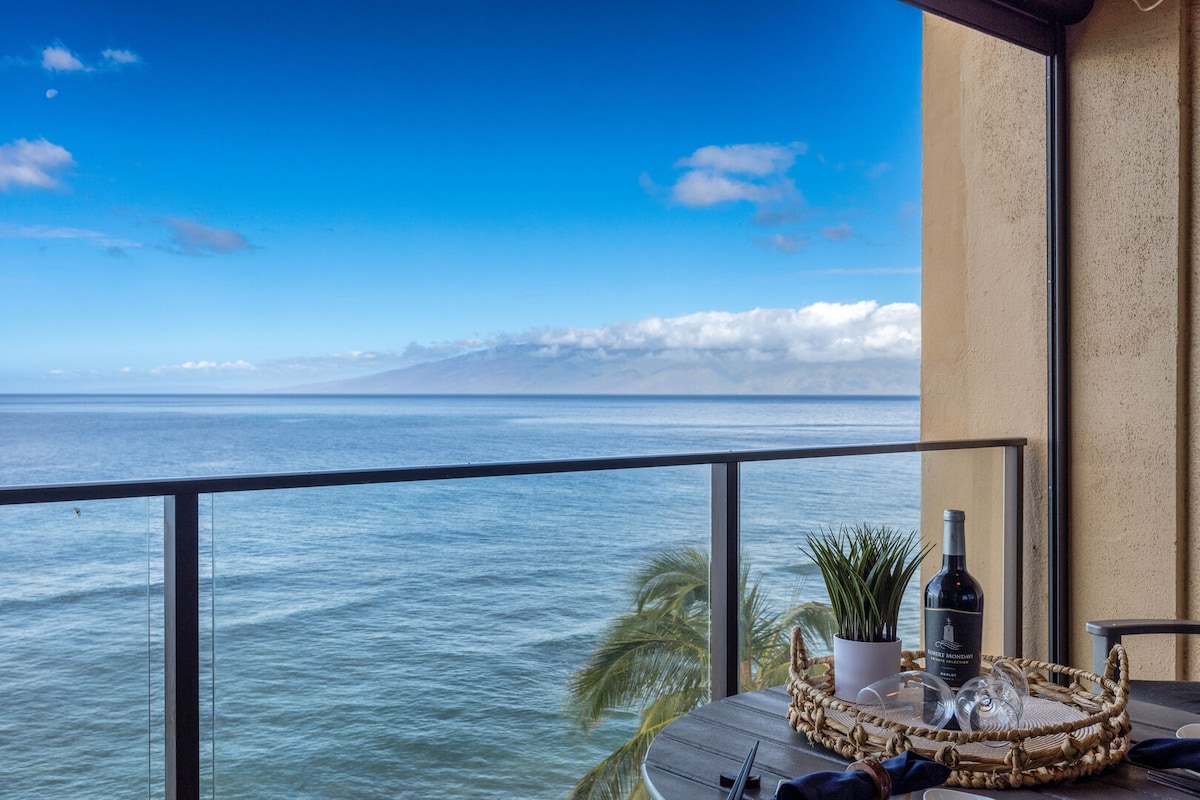 An ocean view is presented from the lanai, featuring a round dining table with a bottle of wine and glasses. Lush greenery is positioned next to the table, and the horizon displays a clear blue sky with scattered clouds over the tranquil water.