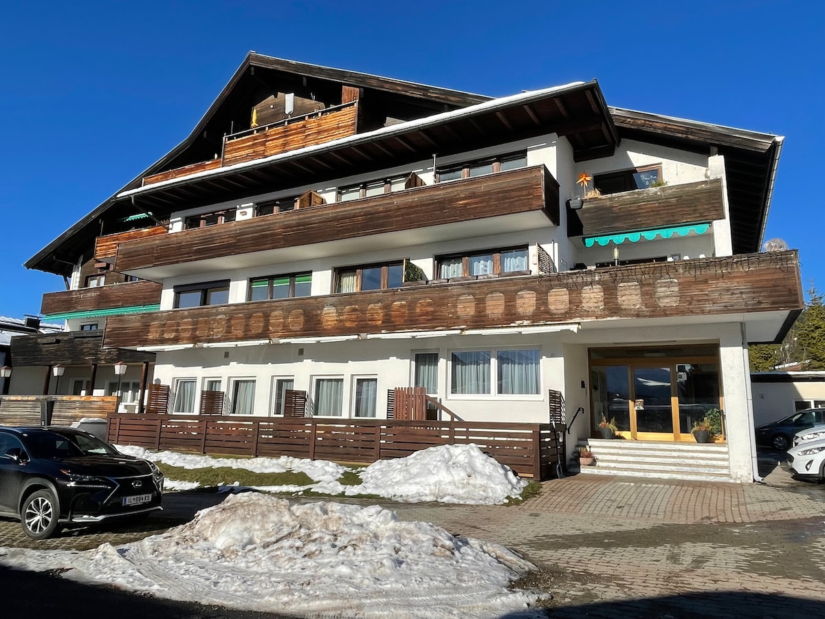 A multi-level building is shown with a combination of wooden and white facades. The upper balconies are adorned with greenery, while snow-covered areas are visible at the base. A parking area in the foreground accommodates vehicles, set against a clear blue sky.
