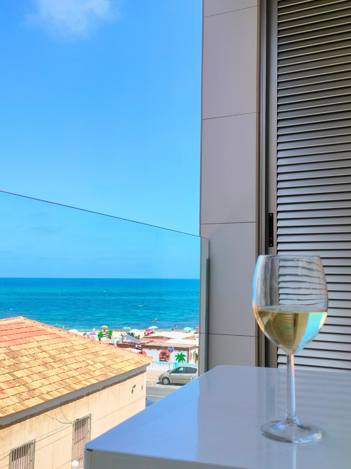 A glass of white wine is placed on a table, overlooking a vibrant blue sea. The view includes a distant beach with sunbathers and colorful umbrellas, while a house with a terracotta roof is visible below. Sunlight brightens the scene, suggesting a warm coastal ambiance.