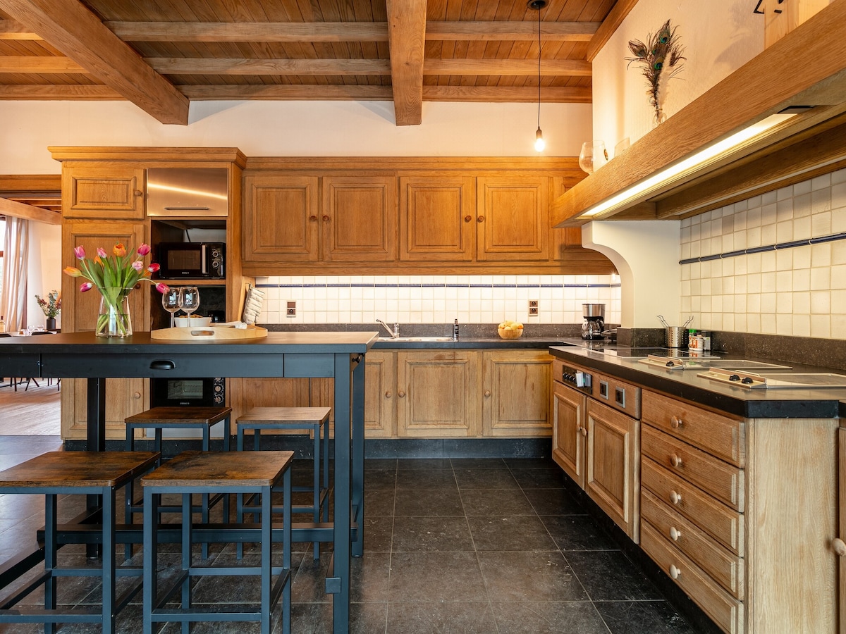 A spacious kitchen features wooden cabinetry and black countertops, providing a welcoming cooking environment. A dining set with tall stools offers seating. Natural light illuminates the space, highlighting the tile floor and wooden beams overhead.